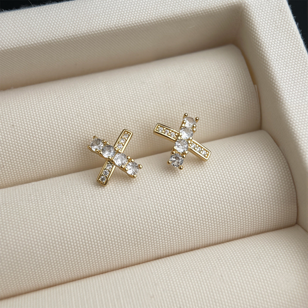 Gold cross-shaped earrings with clear stones on a beige fabric surface